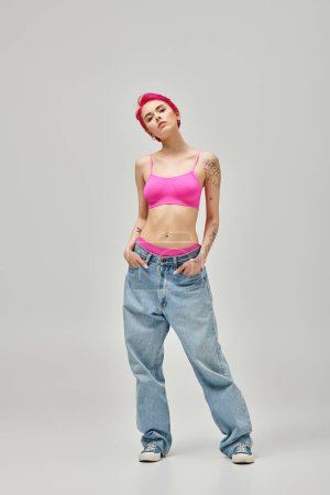 Foto de Mujer atractiva con el pelo corto de color rosa y tatuajes en la parte superior de la cosecha de color rosa y jeans con las manos en los bolsillos - Imagen libre de derechos