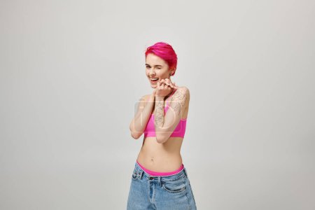 aufgeregte junge Frau mit pinkfarbenen kurzen Haaren posiert in bauchfreiem Top und Jeans auf grauem Hintergrund