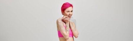 excited young woman with pink short hair posing in crop top and winking on grey backdrop, banner