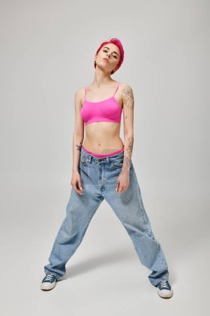 full length, confident woman with pink short hair posing in crop top and jeans on grey backdrop