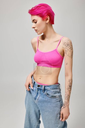 Photo for Confident and tattooed young woman with pink hair posing in crop top and jeans on grey background - Royalty Free Image