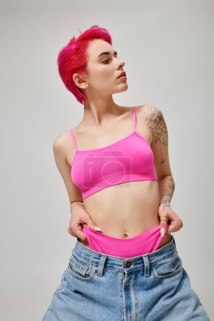 tattooed stylish woman with pink hair posing in crop top and pulling panties from jeans on grey