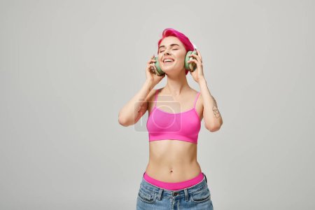 glückliche junge Frau mit rosa Haaren, die Musik in grünen drahtlosen Kopfhörern auf grauem Hintergrund hört