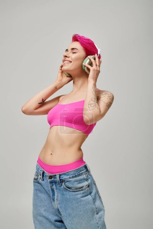 Foto de Mujer elegante tatuada con pelo rosa escuchando música en auriculares inalámbricos sobre fondo gris - Imagen libre de derechos
