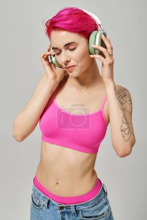 Tätowierte junge Frau mit pinkfarbenen Haaren hört Musik in drahtlosen Kopfhörern vor grauem Hintergrund
