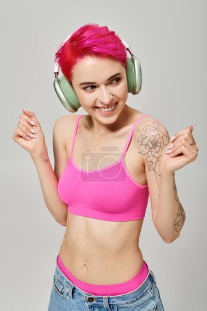 fröhliche tätowierte Frau mit pinkfarbenen Haaren, die in drahtlosen Kopfhörern vor grauem Hintergrund Musik hört