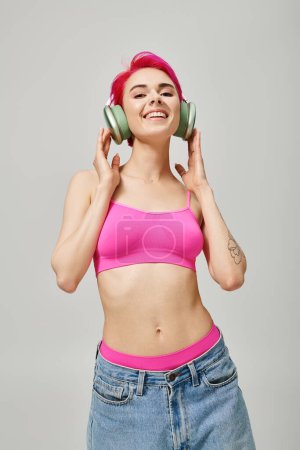 fröhliche gepiercte Frau mit pinkfarbenen Haaren, die in drahtlosen Kopfhörern vor grauem Hintergrund Musik hört