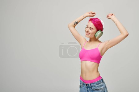 fröhliche und gepiercte Frau mit pinkfarbenen Haaren, die in drahtlosen Kopfhörern vor grauem Hintergrund Musik hört