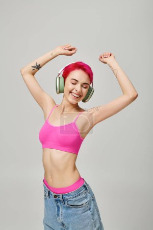 fröhliche und gepiercte Frau mit pinkfarbenen Haaren, die in drahtlosen Kopfhörern vor grauem Hintergrund Musik hört