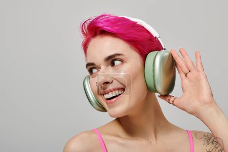 Porträt einer fröhlichen tätowierten Frau mit rosa Haaren, die Musik in drahtlosen Kopfhörern auf grau hört