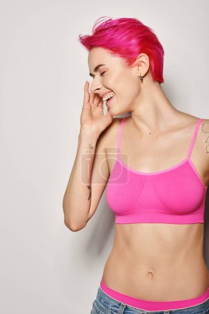 Photo for Side view of tattooed cheerful woman with pink hair posing in crop top and jeans on grey backdrop - Royalty Free Image