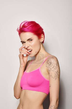 mujer joven tatuada emocional con el pelo rosa posando en la parte superior de la cosecha y uñas mordedoras en el fondo gris