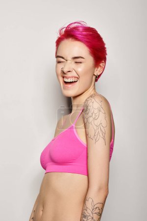 Photo for Portrait of tattooed and excited woman with pink hair laughing with closed eyes on grey backdrop - Royalty Free Image