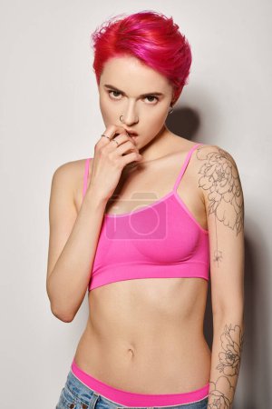 tattooed and pierced woman with pink hair and posing in bright crop top and jeans on grey background