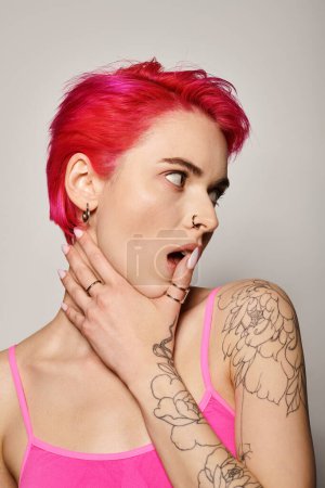tattooed and pierced woman with pink hair and rings on her fingers looking away on grey background