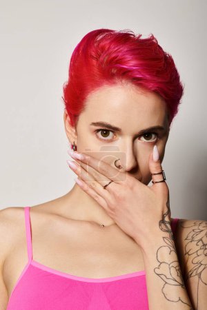 expresiva mujer joven con el pelo rosa cubriendo la boca y mirando a la cámara en el fondo gris