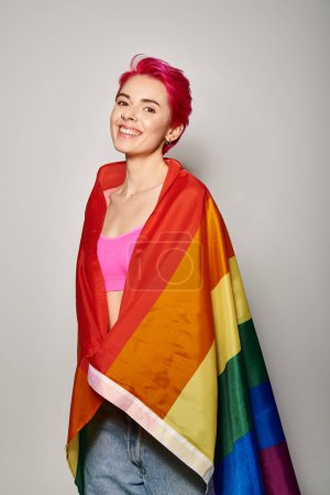 portrait of cheerful young woman with pink hair posing with lgbt rainbow flag on grey backdrop