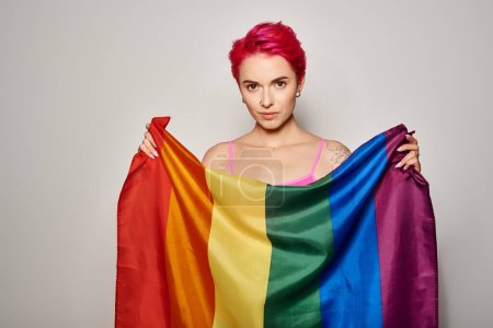 portrait of tattooed young woman with pink hair posing with lgbt rainbow flag on grey backdrop