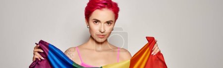 Foto de Retrato de mujer joven tatuada con el pelo rosa posando con la bandera del arco iris lgbt en gris, bandera - Imagen libre de derechos