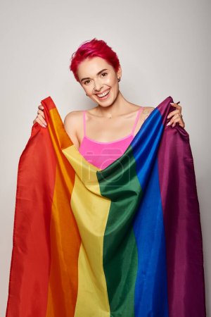 portrait of happy and young woman with pink hair posing with lgbt rainbow flag on grey backdrop