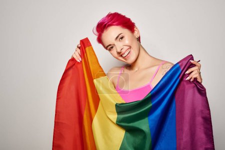 portrait of smiling and young woman with pink hair posing with lgbt rainbow flag on grey backdrop