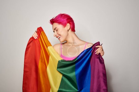 retrato de mujer joven y complacida con el pelo rosa posando con bandera de arco iris lgbt sobre fondo gris