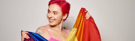 retrato de activista mujer complacida con el pelo rosa posando con la bandera del arco iris lgbt en gris, bandera