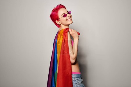 joven y complacida activista femenina con pelo rosa y gafas de sol posando con bandera de arco iris lgbt