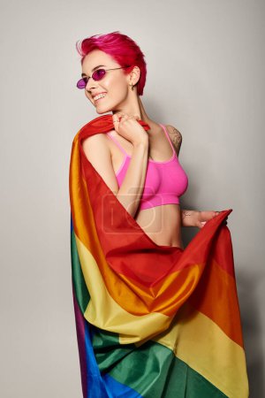 joven y feliz activista femenina con pelo rosa y gafas de sol posando con bandera de arco iris lgbt sobre gris