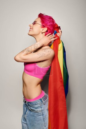 mujer joven y alegre con pelo rosa y gafas de sol posando con bandera de arco iris lgbt sobre fondo gris