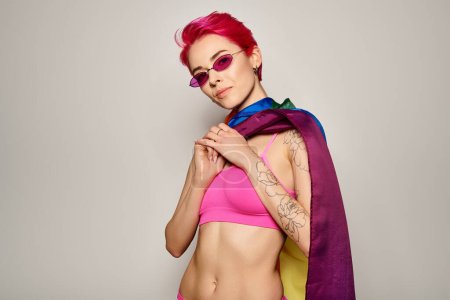 young woman with pink hair and stylish sunglasses posing with lgbt rainbow flag on grey backdrop