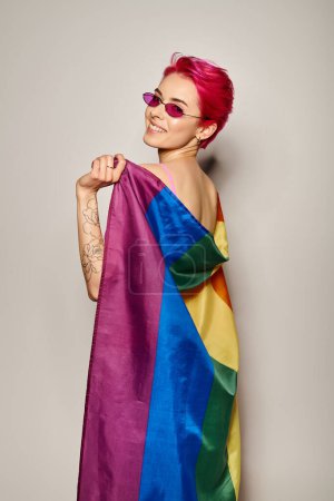 Foto de Joven activista femenina feliz con pelo rosa y gafas de sol posando con bandera de arco iris lgbt en gris - Imagen libre de derechos