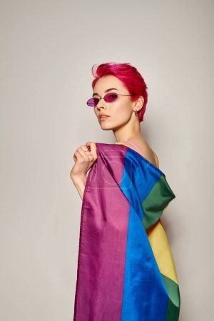 mujer joven y confiada con pelo rosa y gafas de sol posando con bandera de arco iris lgbt sobre fondo gris