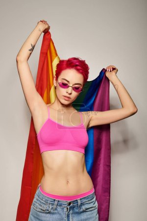 Foto de Mujer joven tatuada con pelo rosa y gafas de sol posando con bandera de arco iris lgbt sobre fondo gris - Imagen libre de derechos