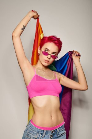 mujer joven tatuada con pelo rosa y gafas de sol posando con bandera de arco iris sobre fondo gris