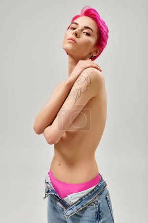 mujer joven tatuada y en topless con el pelo rosa cubriendo los pechos y posando sobre fondo gris