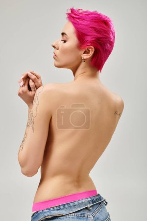 Rückseite, tätowierte und oben ohne junge Frau mit pinkfarbenen kurzen Haaren posiert auf grauem Hintergrund