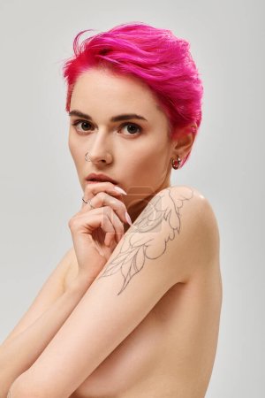 retrato de mujer joven tatuada y en topless con el pelo rosa cubriendo los pechos en el fondo gris