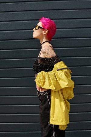 Téléchargez les photos : Vue latérale de la jeune femme aux cheveux roses en lunettes de soleil et tenue tendance posant à l'extérieur, style rue - en image libre de droit