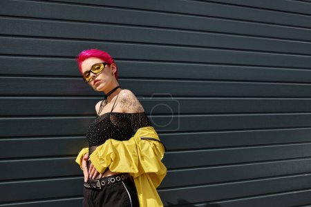 jeune femme aux cheveux roses en lunettes de soleil jaunes et tenue élégante posant à l'extérieur, style rue
