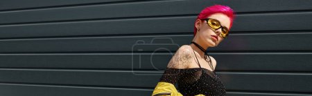 pink haired young woman in yellow sunglasses and stylish outfit posing outdoors, street banner