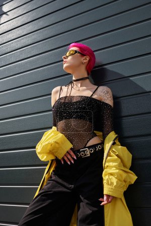 rosa haarige junge Frau mit gelber Sonnenbrille und stylischem Outfit posiert im Freien, Blick aus dem niedrigen Winkel