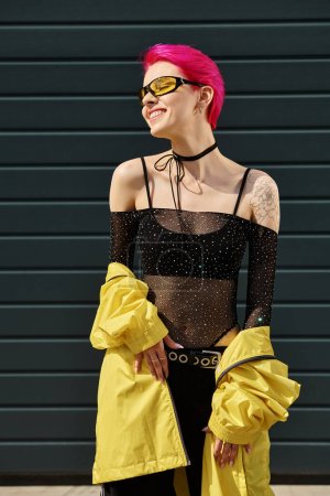 pink haired happy woman in yellow sunglasses and trendy outfit posing on urban street outdoors