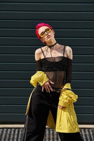 Téléchargez les photos : Jeune femme aux cheveux roses en lunettes de soleil jaunes et tenue élégante posant sur la rue urbaine à l'extérieur - en image libre de droit