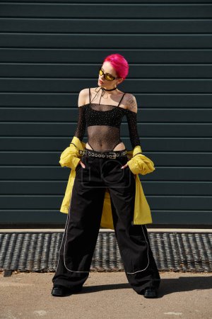 pink haired young woman in sunglasses and stylish attire posing with hands in pockets on street