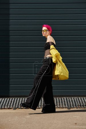 rosa haarige junge Frau mit gelber Sonnenbrille und stylischer Kleidung, die draußen auf der Straße spaziert