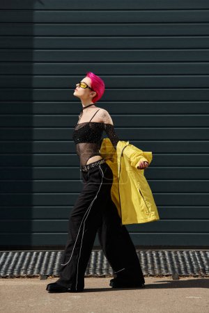 rosa haarige junge Frau mit gelber Sonnenbrille und trendiger Kleidung, die draußen auf der Straße spaziert