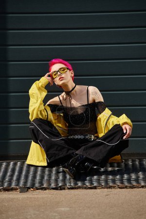 pink haired young woman in yellow sunglasses and trendy attire sitting on urban street outdoors