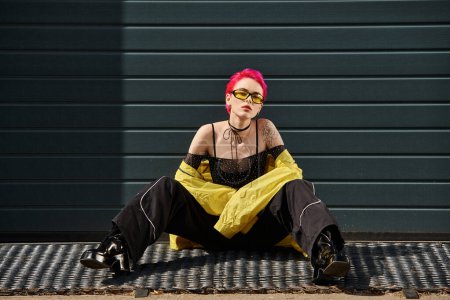 young woman with pink hair and tattoo posing in yellow sunglasses and stylish attire on urban street