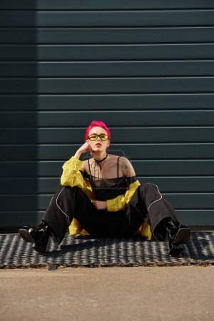 jeune femme aux cheveux roses et tatouage posant en lunettes de soleil jaunes et tenue tendance dans la rue urbaine
